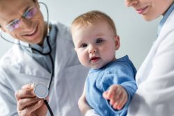 Doctor with a stethoscope getting ready to listen to baby's heart while baby looks at and points to the camera. 