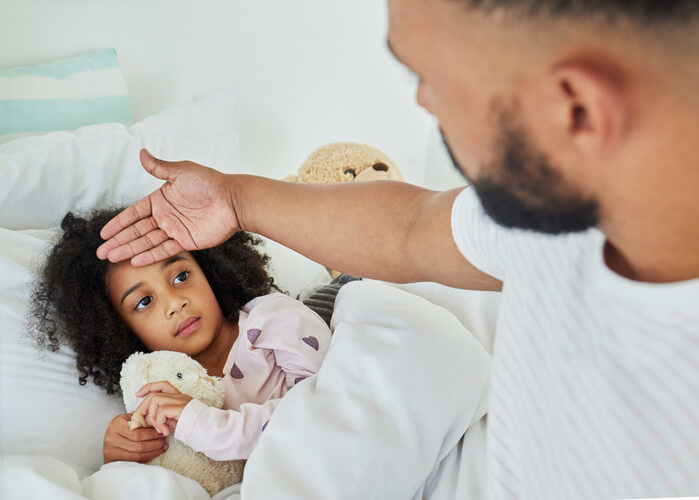 Child lays in bed while her parent checks her forehead for a fever.
