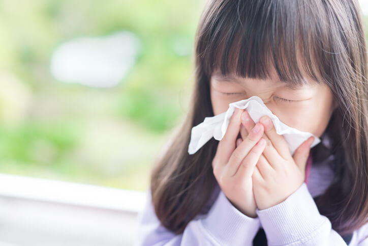A sick child blows her nose, holding a tissue to her face.
