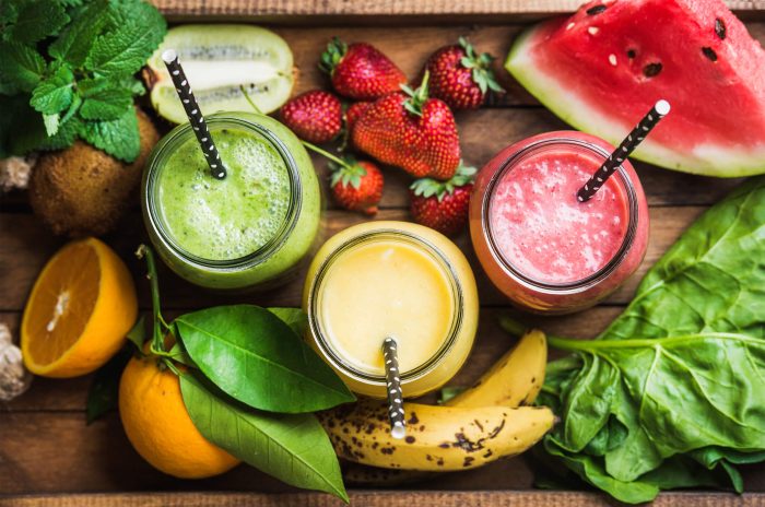 Three smoothies are depicted, surrounding by fruits and vegetables. Smoothies are a great soft food for illness recovery.